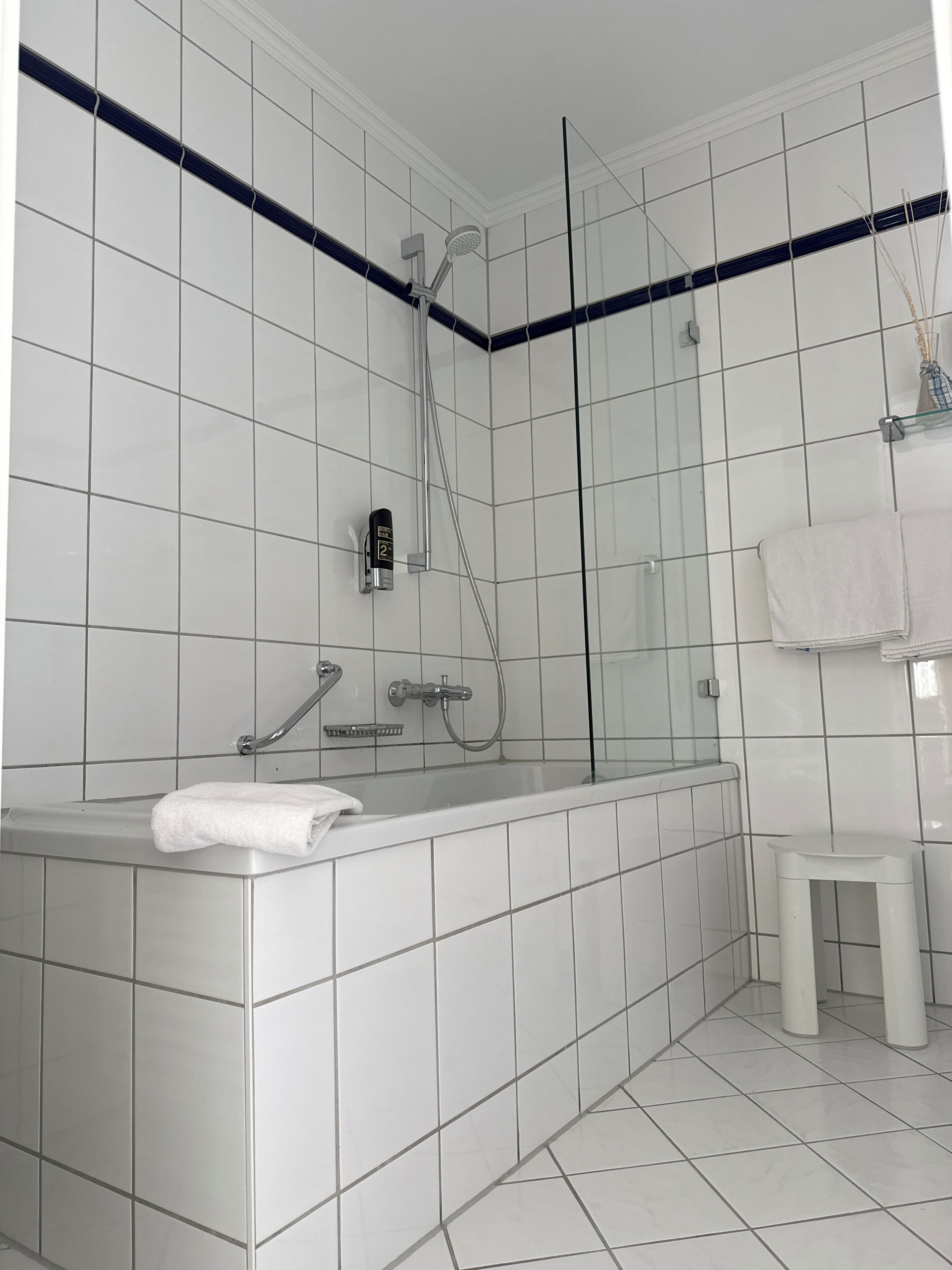 Un baño con paredes de azulejos blancos, cerramiento de ducha de cristal, una bañera blanca y un taburete pequeño blanco.