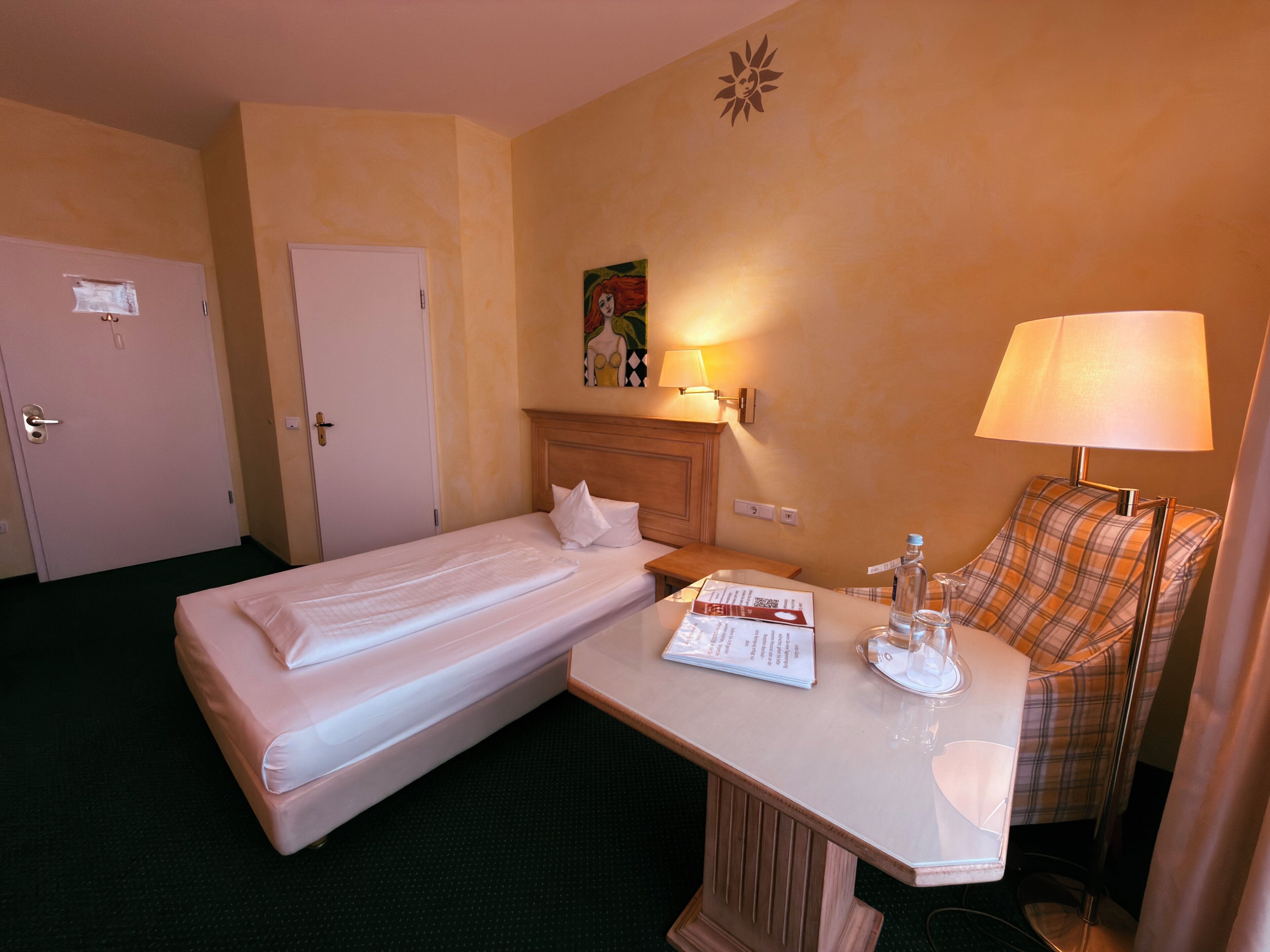 A hotel room with a single bed, a small table with a lamp, a chair, and a painting on the wall.