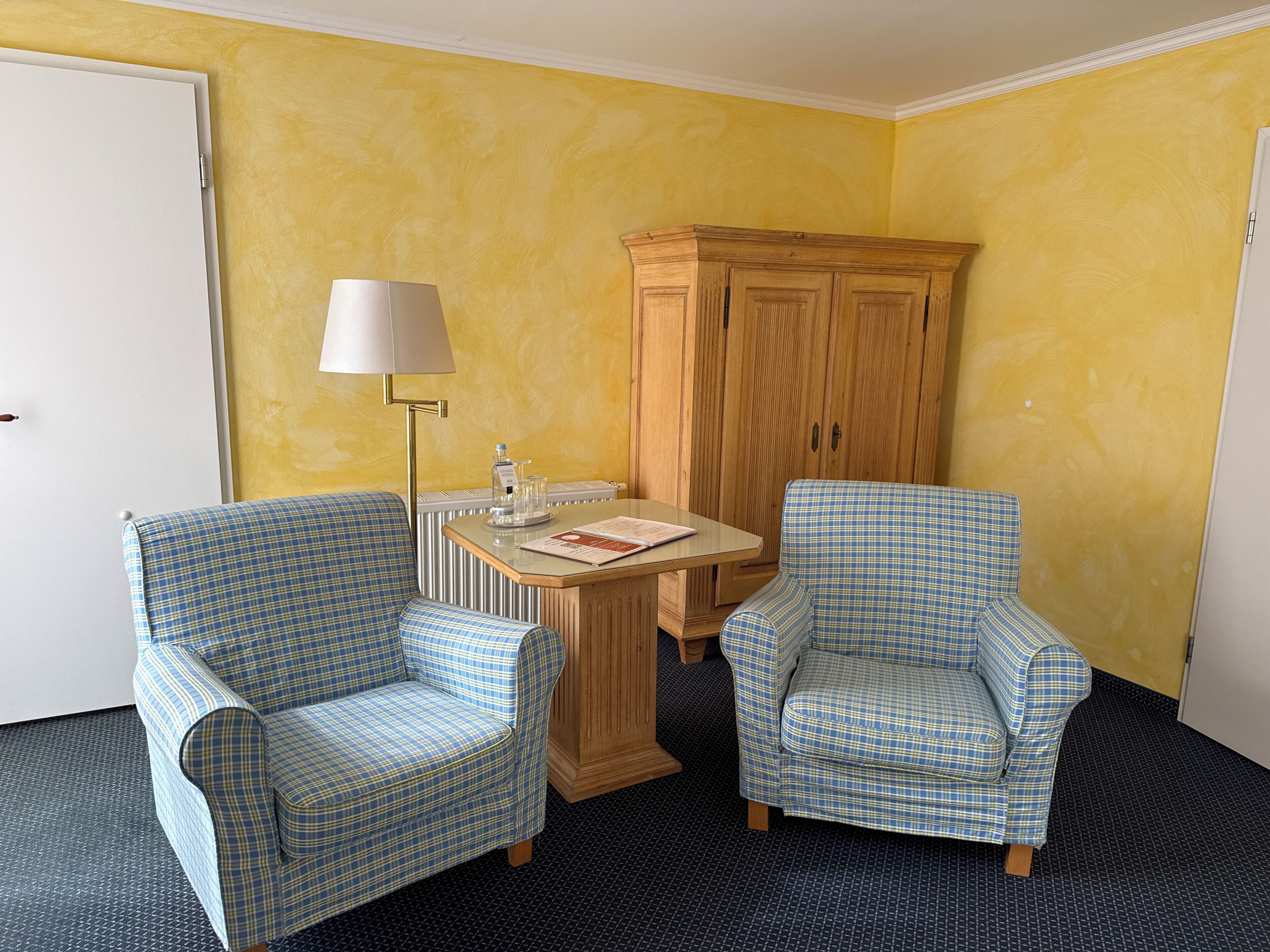 A room with two checkered armchairs, a small table with a lamp, a wooden cabinet, and a door.