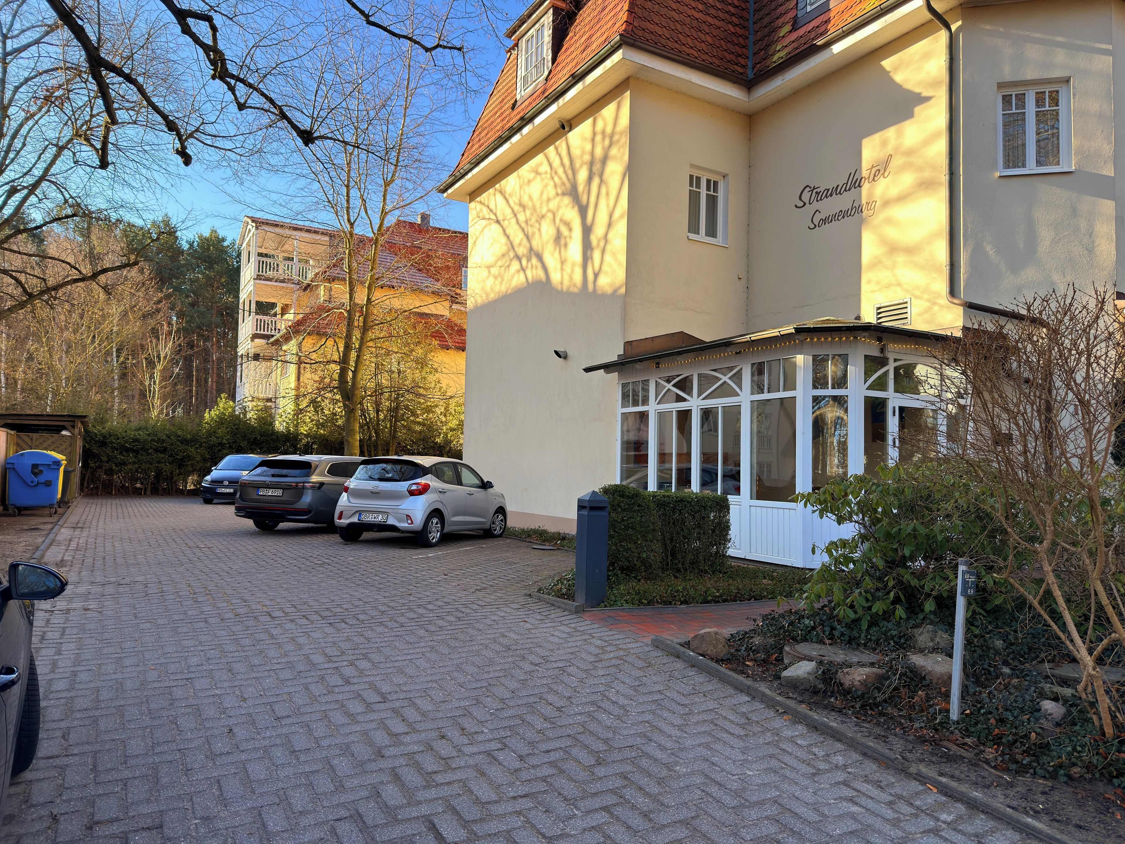 A building with a sign reading 'Strandhotel Soverburg,' a paved driveway, parked cars, and a small garden area.