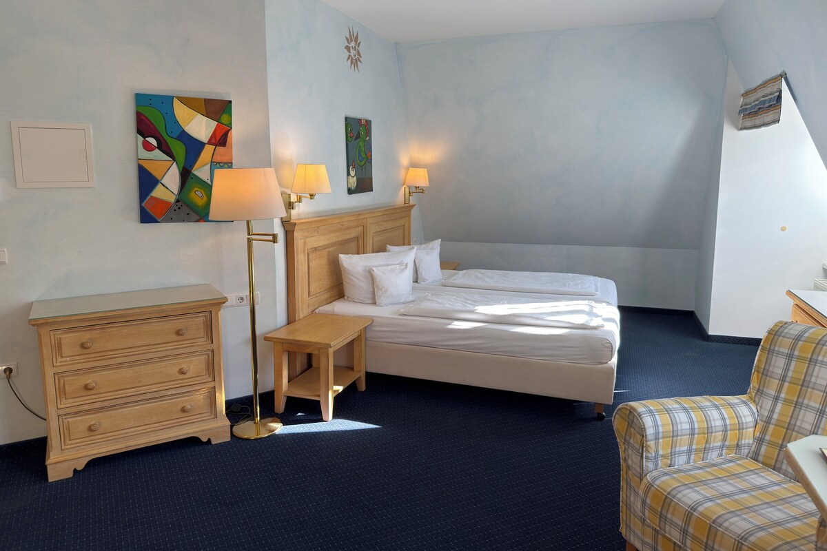 A hotel room with a bed, bedside tables, a chair, and artwork on the walls.