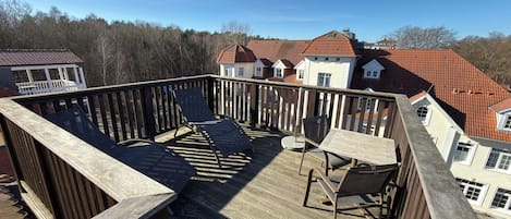 Une terrasse en bois avec des meubles d’extérieur, une vue sur des maisons aux toits rouges et un ciel dégagé.