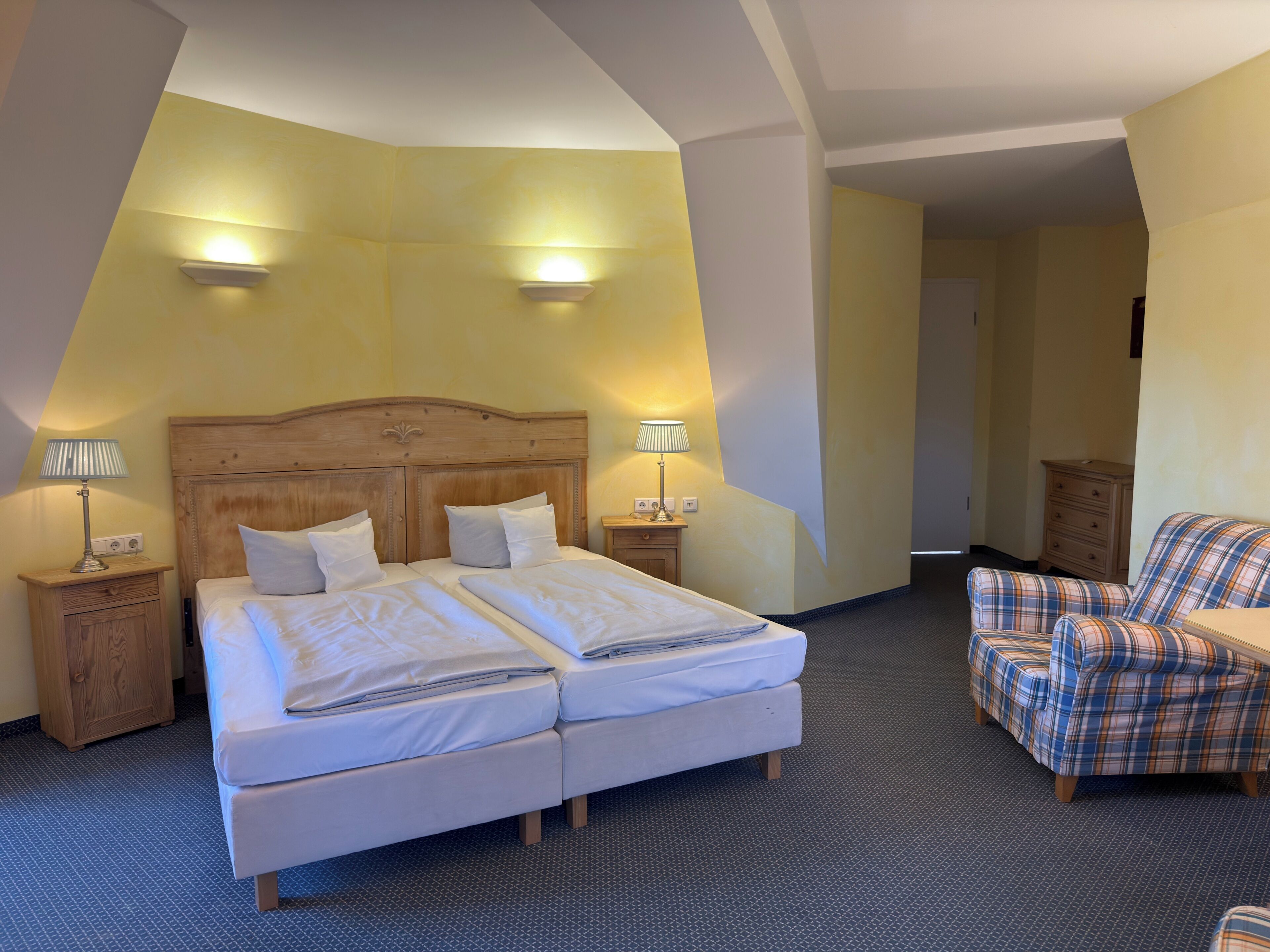 A hotel room with a double bed, two bedside tables with lamps, a plaid armchair, and a small wooden dresser.