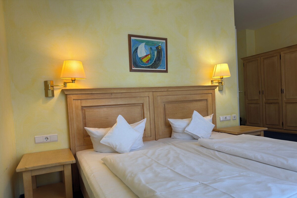 A hotel room with a double bed, two bedside tables, a headboard with a painting, and two wall-mounted lamps.