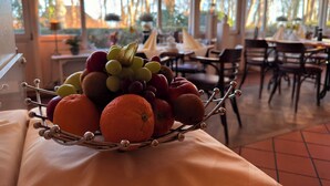 A dining area with a table set for two, a fruit bowl on a table, and a view of trees through large windows. - Strandhotel Sonnenburg by ANS (Kuehlungsborn)