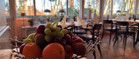 A dining area with a table set for two, a fruit bowl on a table, and a view of trees through large windows.