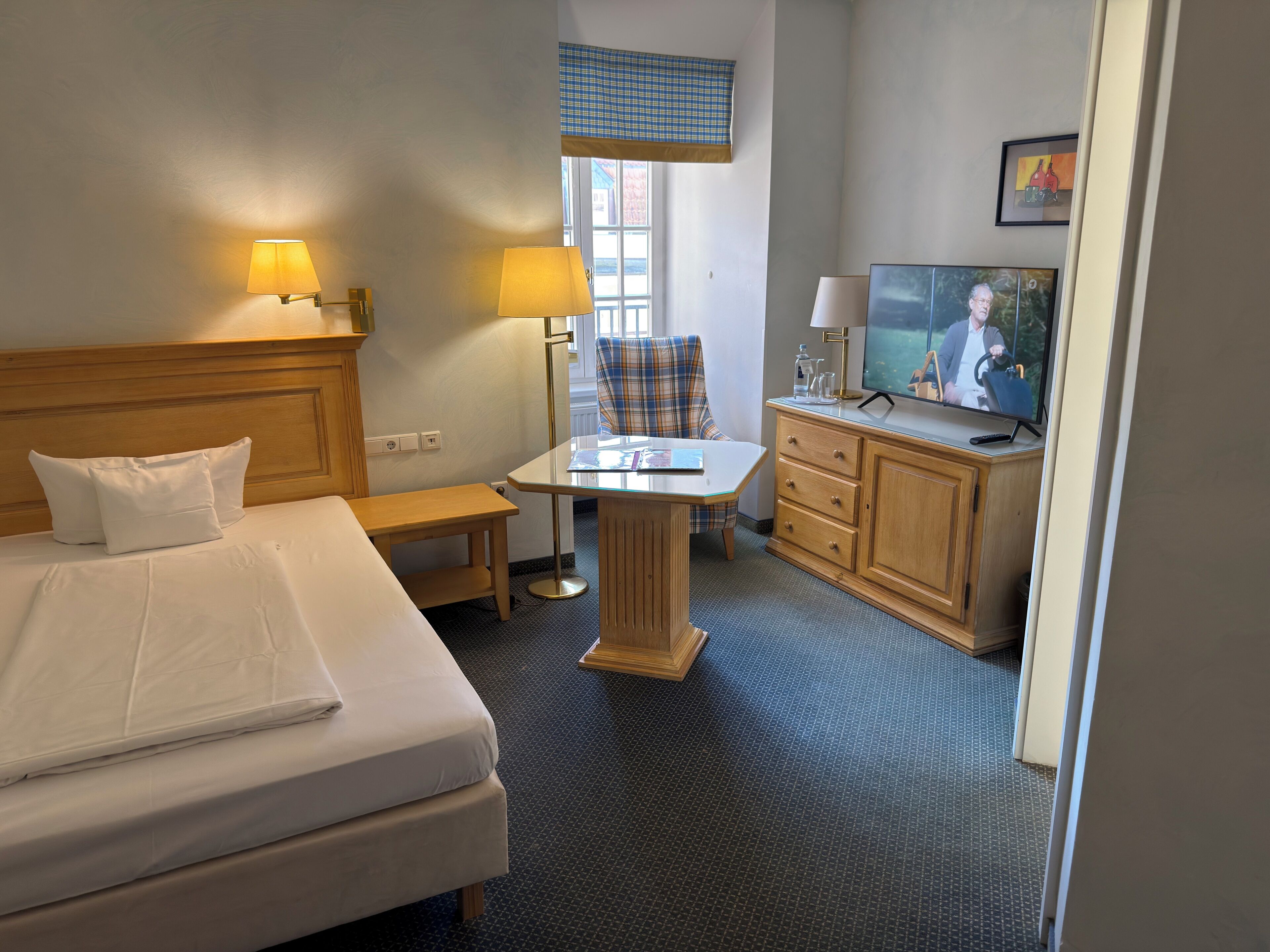 A hotel room with a bed, bedside lamps, a desk with a chair, a television, and a window with a view.