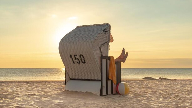 A beach chair with a number '150' on it, positioned on a sandy beach with a view of the ocean and a sunset in the background. - Hotel Wittenbeck by ANS (Wittenbeck)