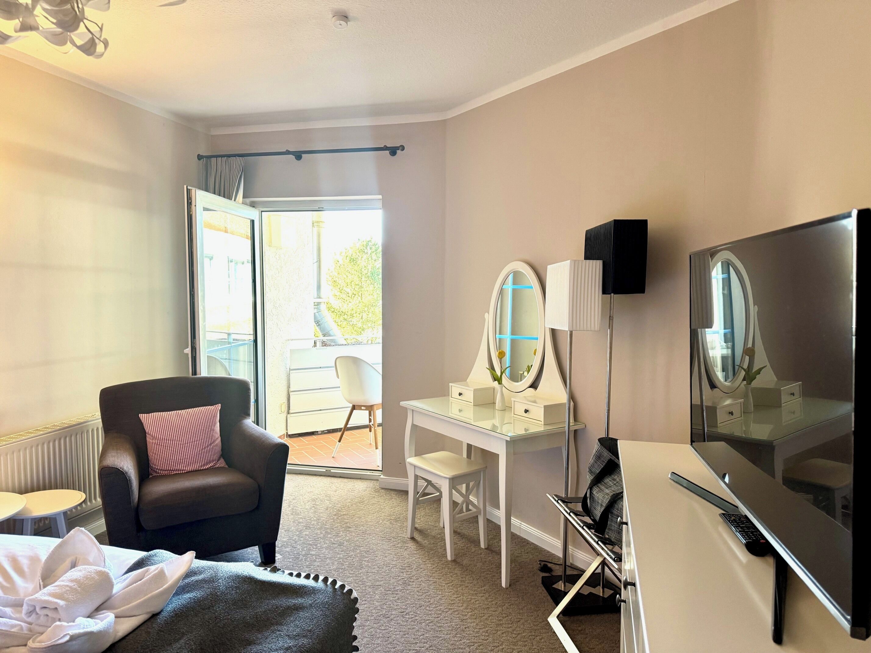A hotel room with a bed, a chair, a vanity with a mirror, a desk, a flat-screen TV, and a window with a view of the outside.