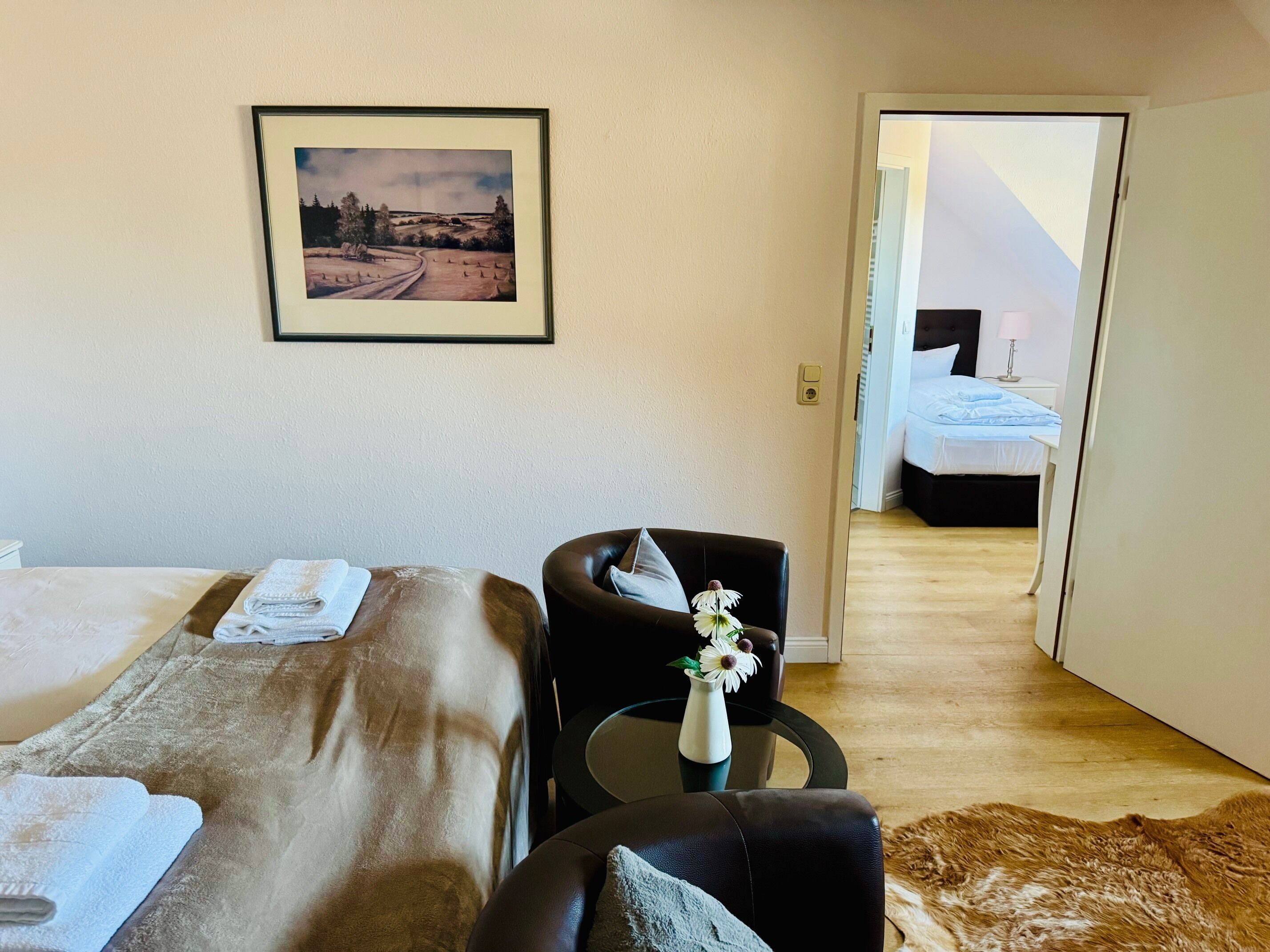 A hotel room with two beds, a desk, a chair, a vase with flowers, and a framed picture on the wall.