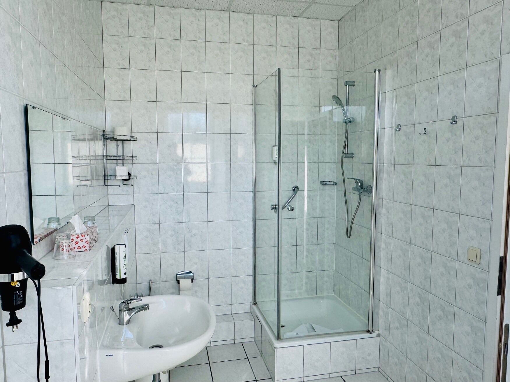 A bathroom with a glass-enclosed shower, a white sink, a mirror, and a towel dispenser.