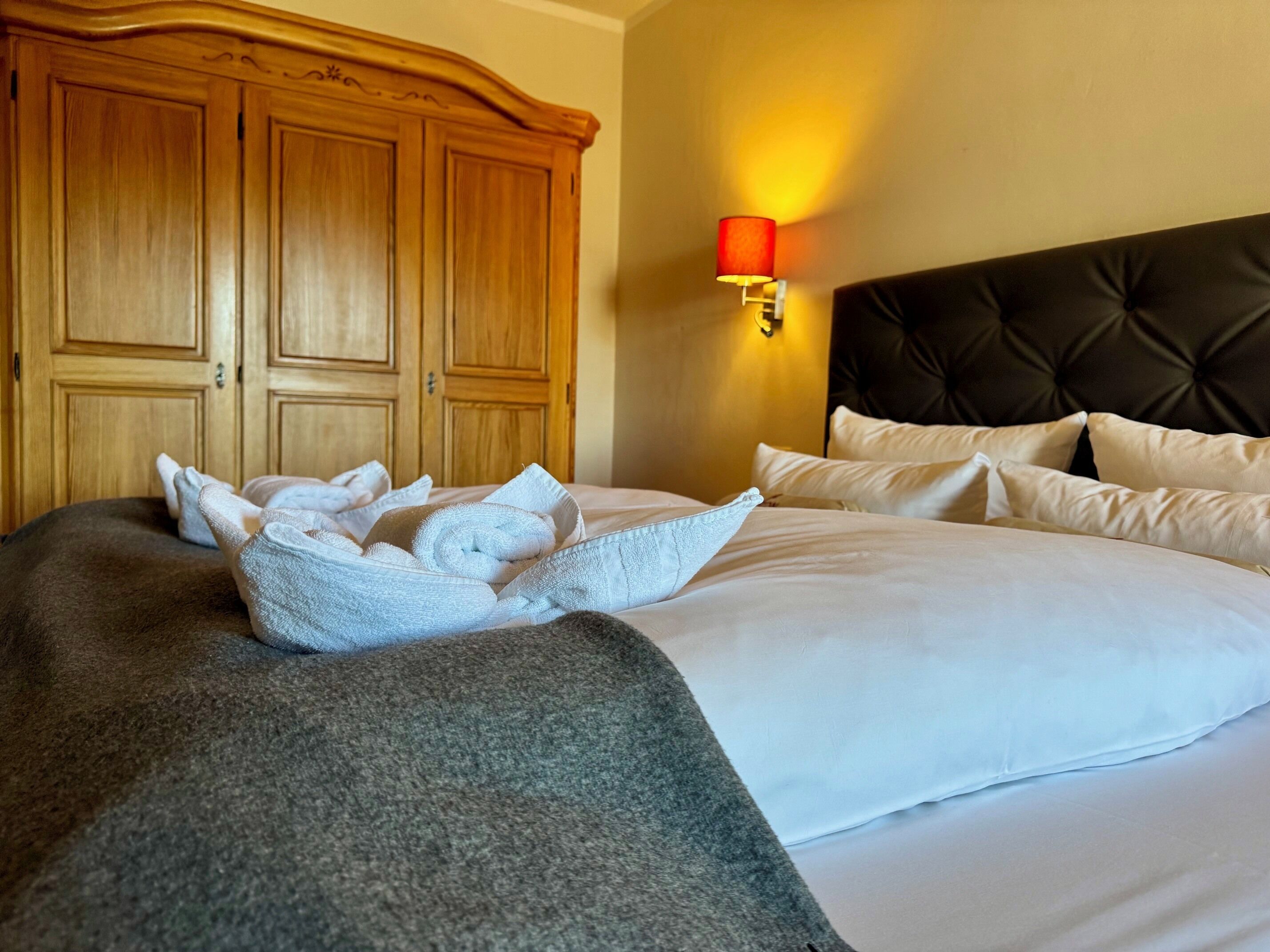 Quarto de hotel com uma cama grande, um guarda-roupa de madeira e uma luminária fixada na parede.