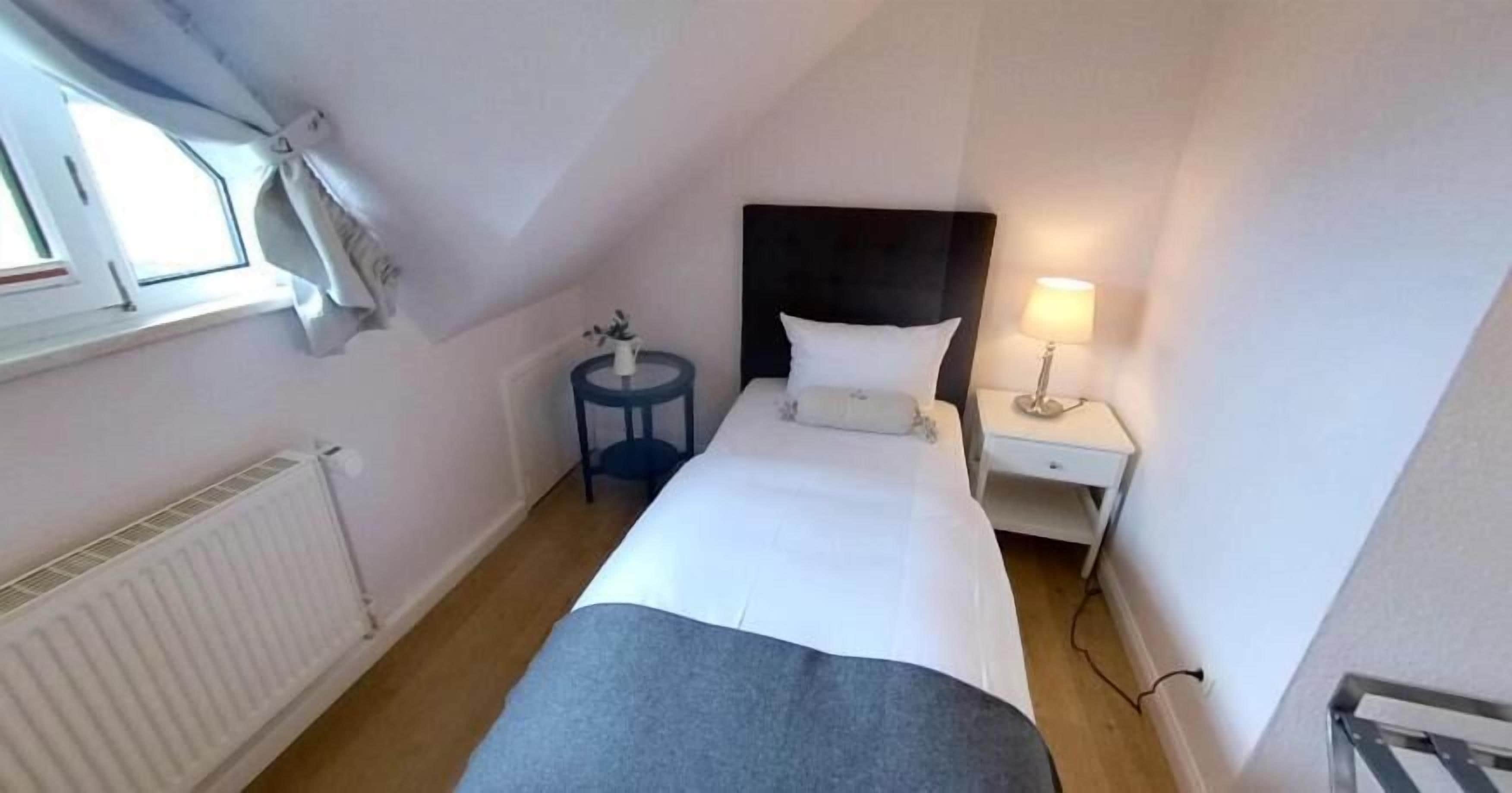 Une petite chambre d’hôtel étroite, avec un lit simple, une table de chevet avec une lampe, une petite table ronde avec une plante et une fenêtre avec des rideaux.