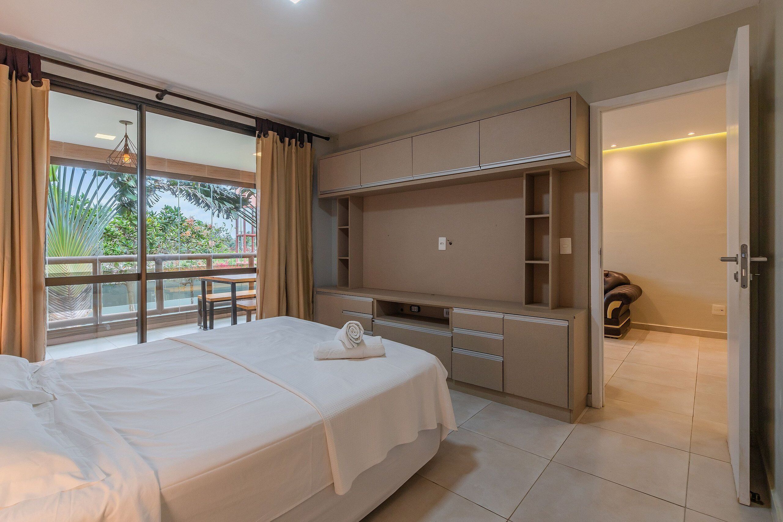 A hotel room with a large bed, a desk with a chair, a wardrobe, and a view of greenery through a sliding door.