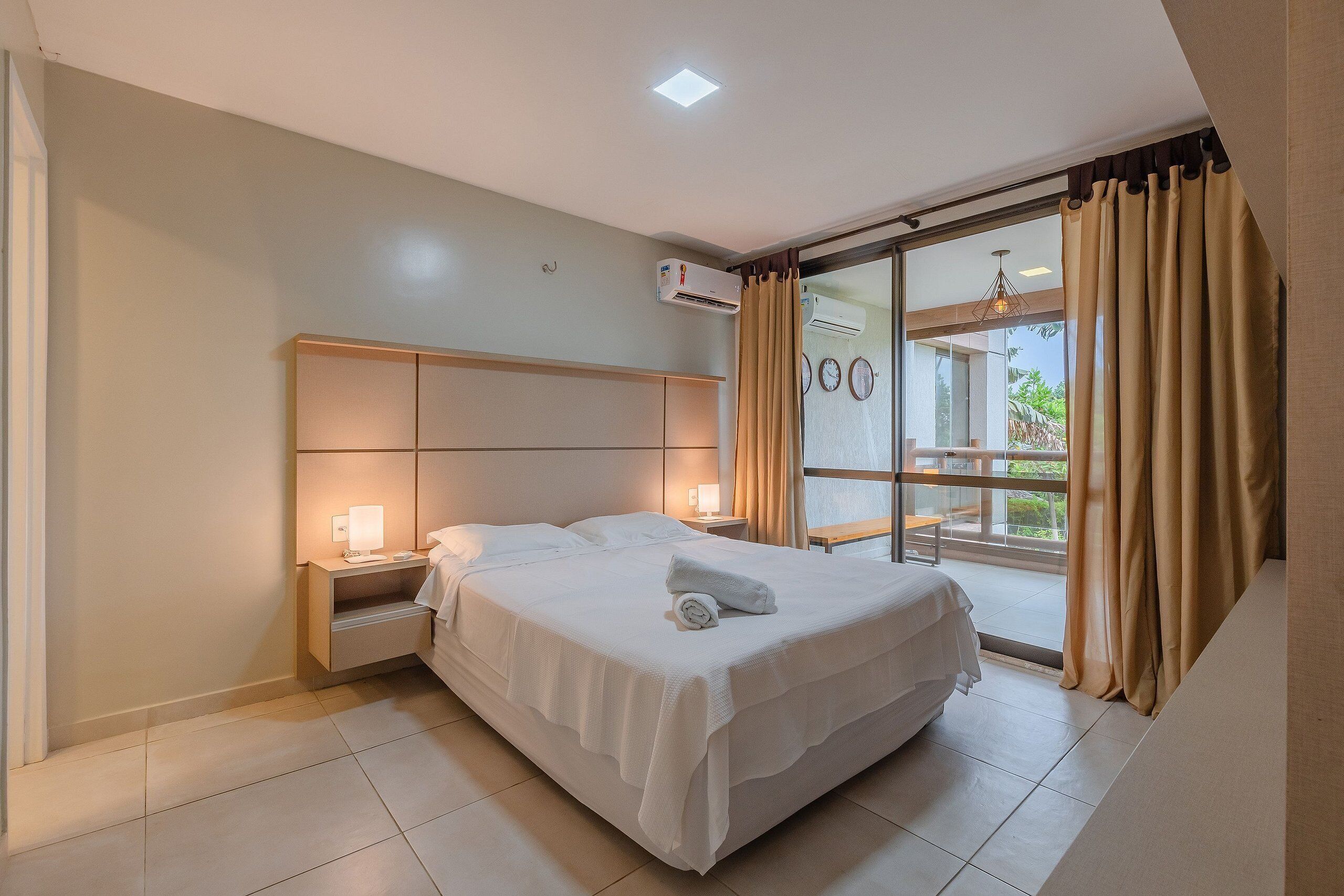A neatly made bed with a white comforter and a rolled towel, a bedside table with a lamp, and a window with curtains.