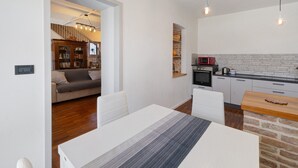 Een moderne keuken met een magnetron, fornuis en een houten aanrecht. Er is een eethoek met een tafel en stoelen, en een trap die leidt naar een woonkamer met een bank en een boekenkast.