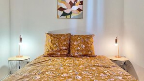 A bedroom with a floral bedspread, two bedside lamps, and a wall-mounted abstract painting. - Quiet apartment near train station and Quimper historic center (Quimper)