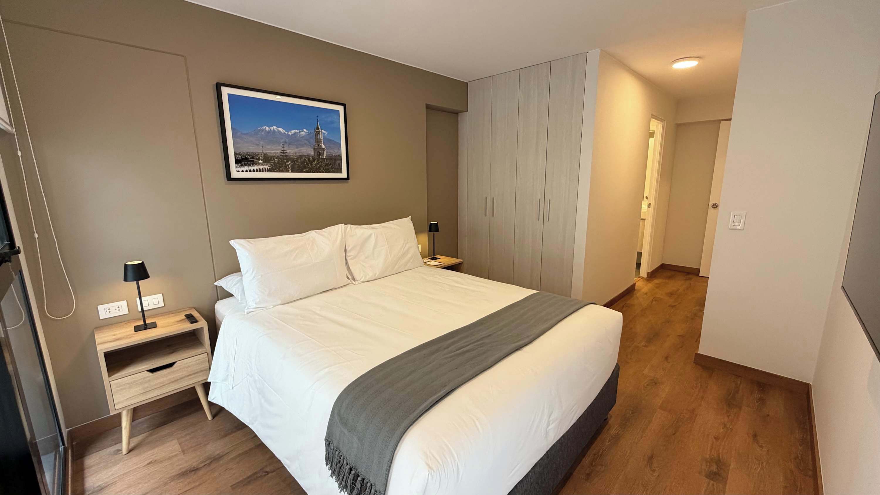 A modern hotel room with a large bed, a nightstand, a lamp, a wardrobe, and a framed picture on the wall.