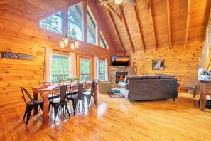 A cabin interior with a dining area, sofa, fireplace, and large windows. - Cozy 1BD+Loft w/ Hot Tub! Fireplace + Pool Access! (Pigeon Forge)