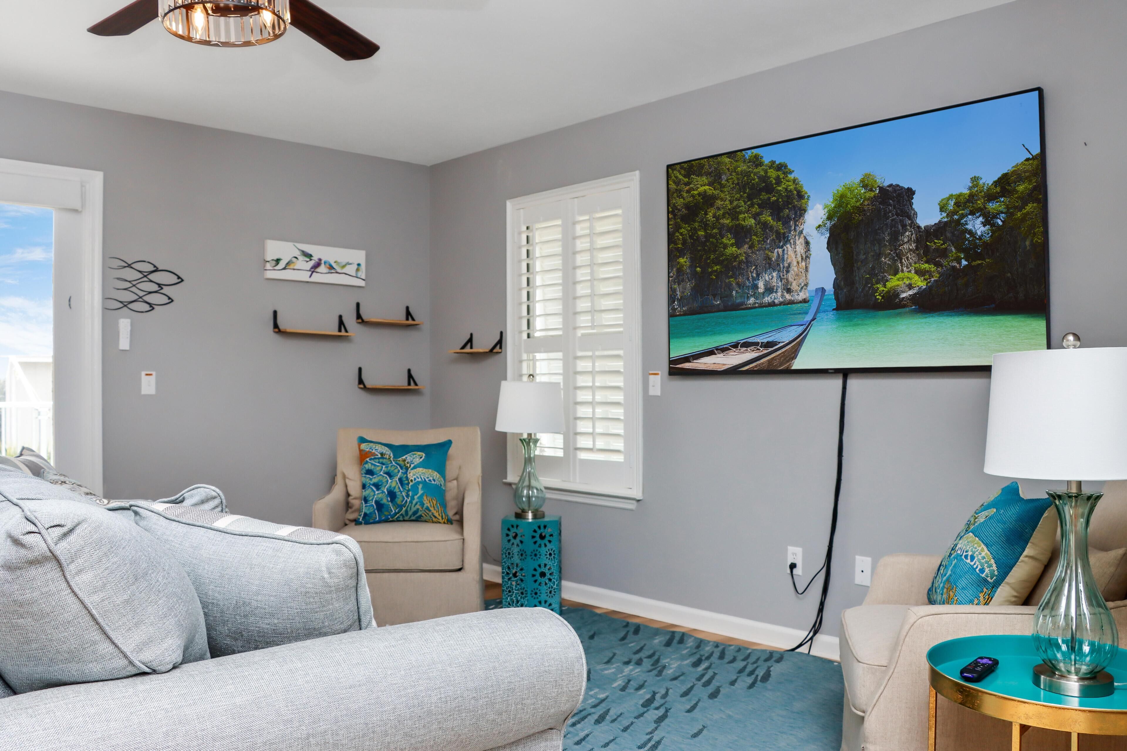 A living room with a grey sofa, a beige armchair, a blue patterned rug, a TV displaying a tropical scene, and a teal side table with a lamp.