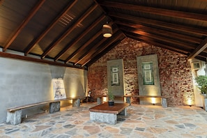 A stone-floored room with a wooden ceiling, benches, a table with a plant, and framed artwork on the walls.