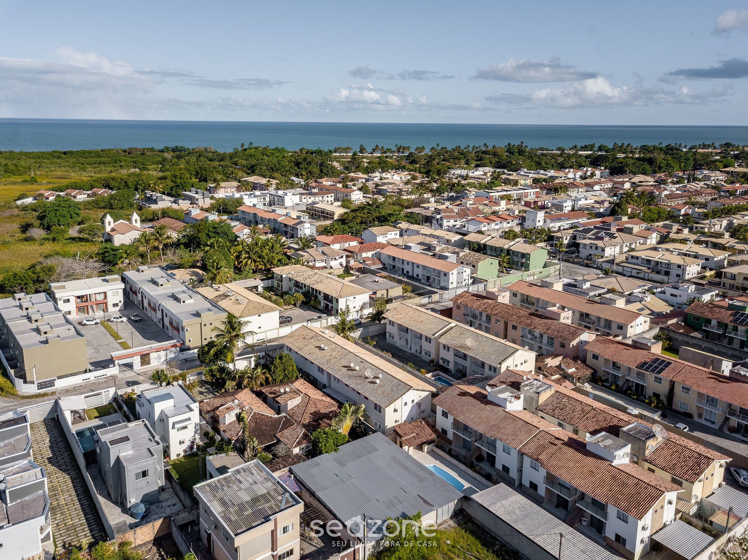 An aerial view of a residential area with houses, some with red-tiled roofs, and a large body of water in the background.