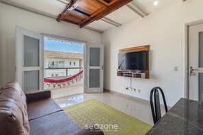 Un salon comprenant un canapé, une table à manger, un téléviseur fixé au mur et une vue sur un bâtiment à l’extérieur.