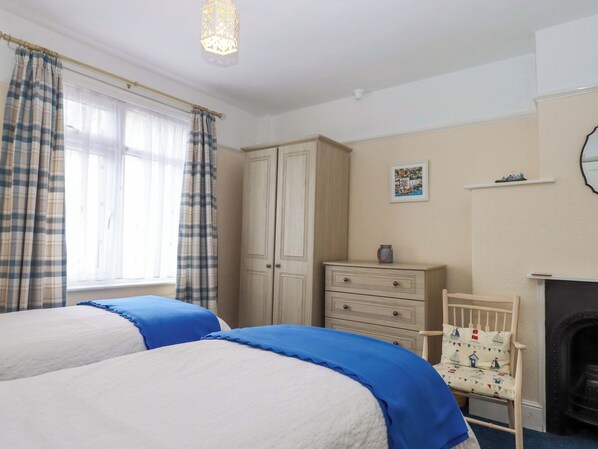 A bedroom with two beds, a dresser, a chair, a fireplace, and a window with curtains. - Elmbank Gardens (Paignton)