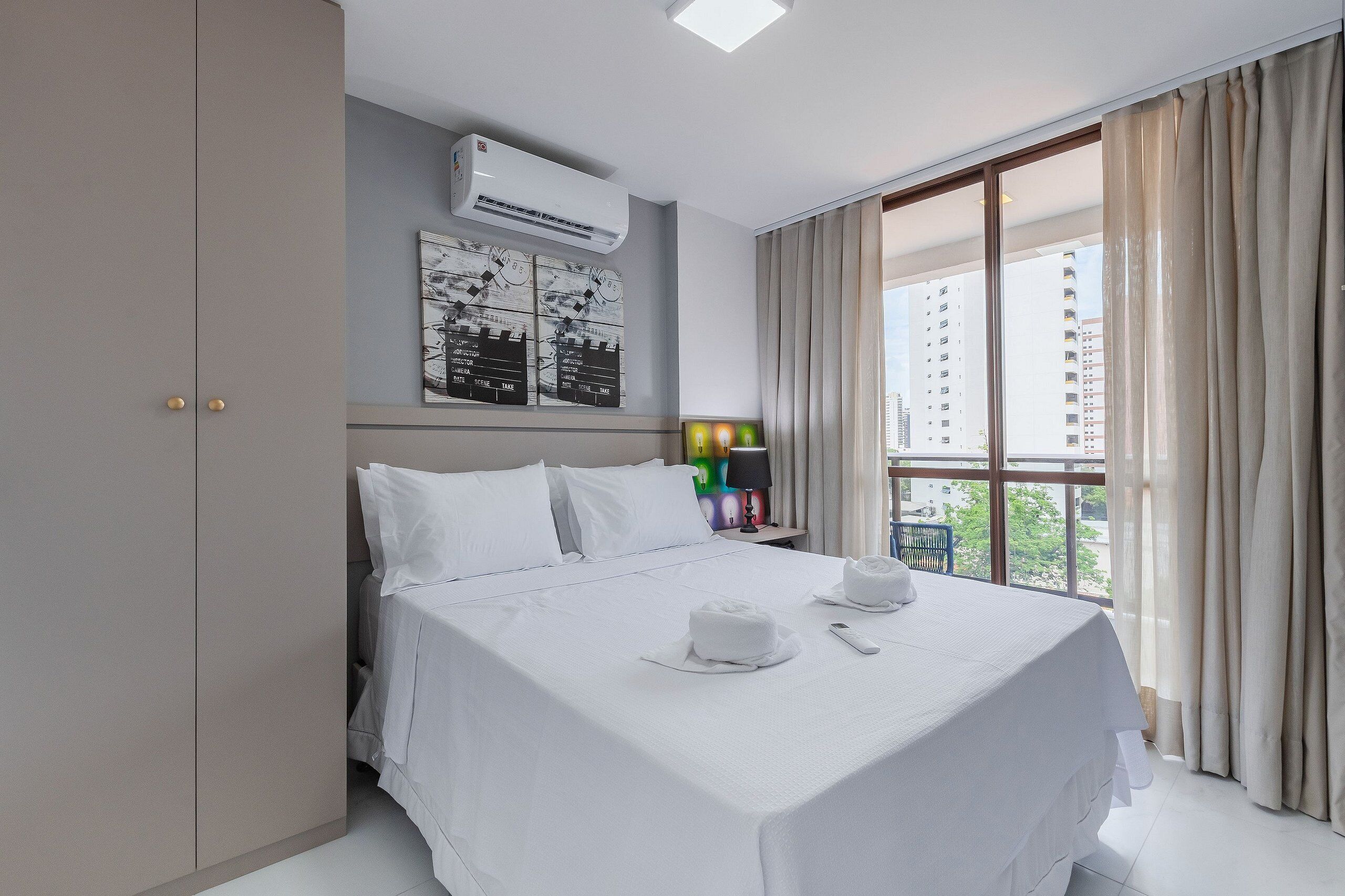 Een hotelkamer met een groot bed, een kledingkast, een airconditioner, een nachtkastje met een lamp en een raam met gordijnen.