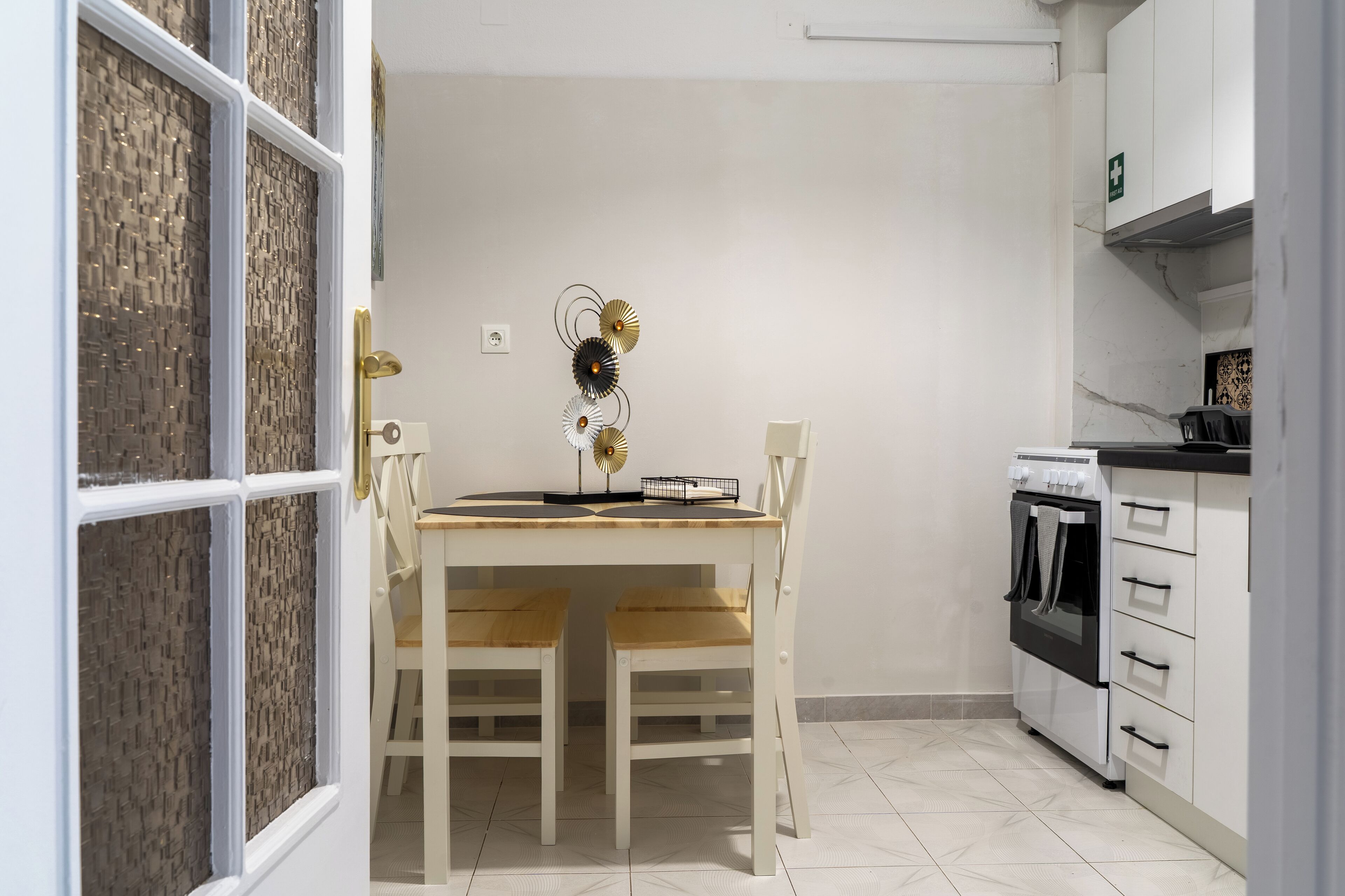 A kitchen with a white stove, a white oven, and a white cabinet with drawers. There is a white table with two chairs and a decorative wall piece above it.