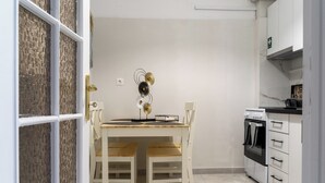 A kitchen with a white stove, a white oven, and a white cabinet with drawers. There is a white table with two chairs and a decorative wall piece above it. - Del Sol 6 Meteora Luxurious Family Suites (Kalampaka)