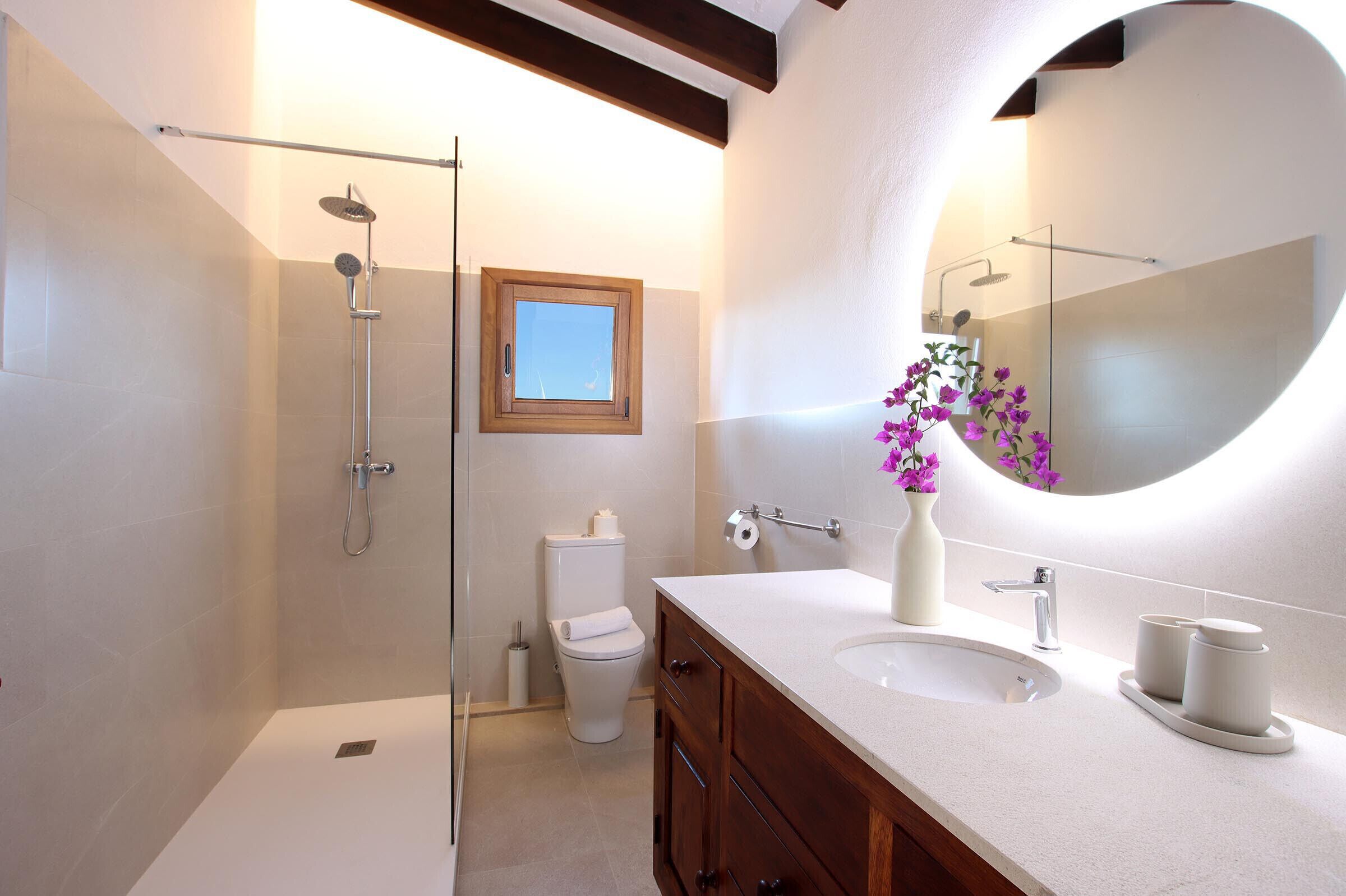 A modern bathroom with a glass-enclosed shower, a toilet, a sink with two cups, and a mirror with a round frame.