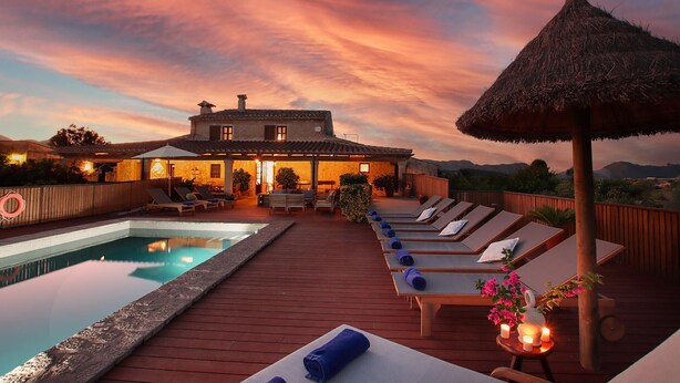 Een zwembadterras met ligbedden, een rieten parasol en een uitzicht op een huis bij zonsondergang.