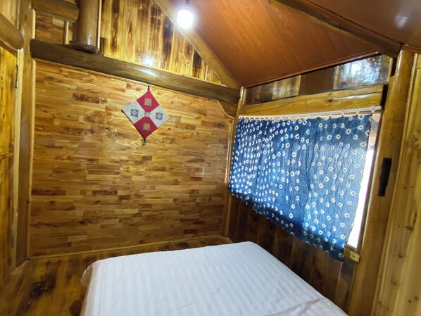 Habitación de cabaña de madera con una cama, un colcha colgada en la pared y una ventana con cortina estampada.