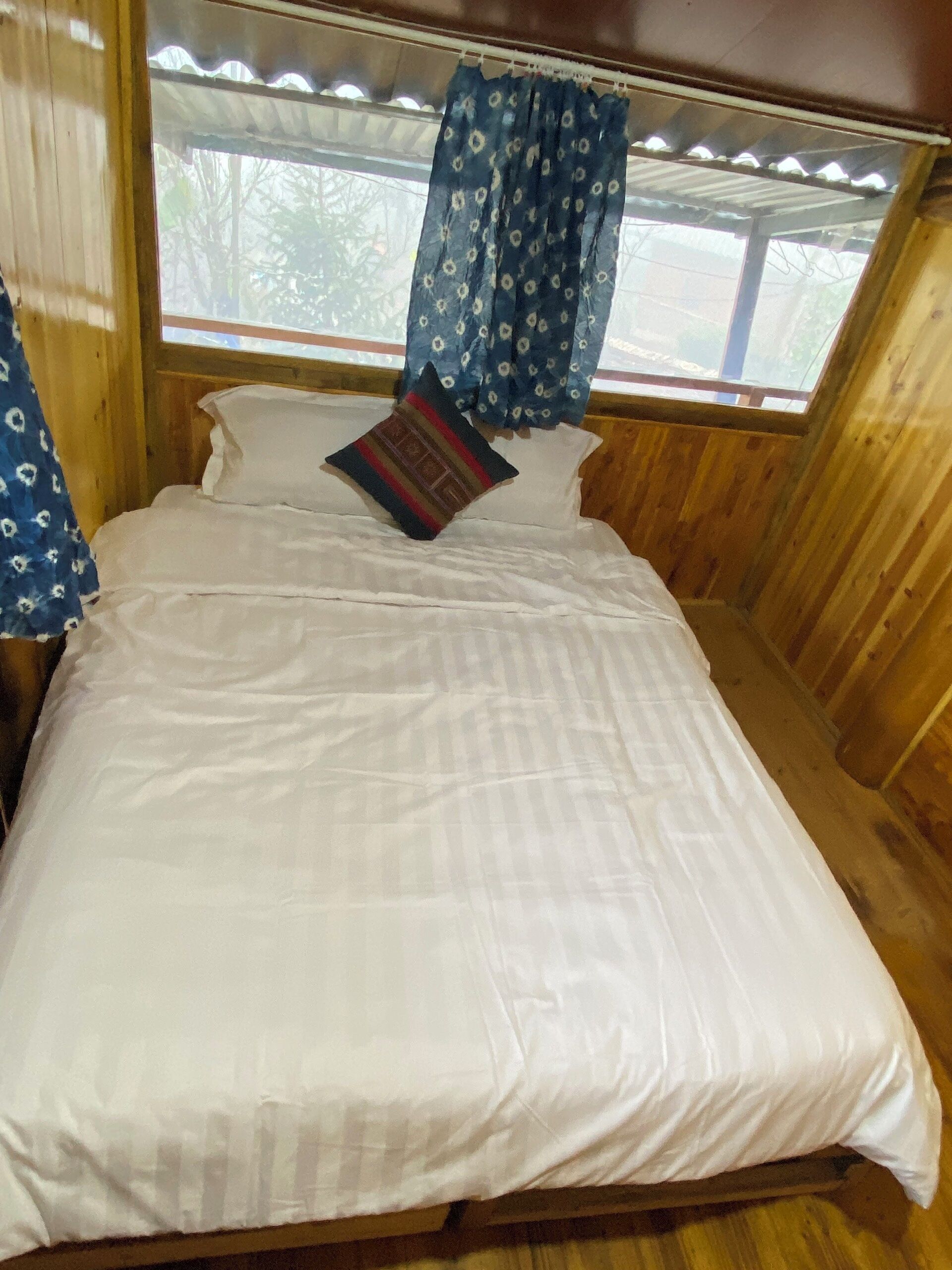 Una stanza in legno con un letto, una finestra con tenda e vista su alberi.
