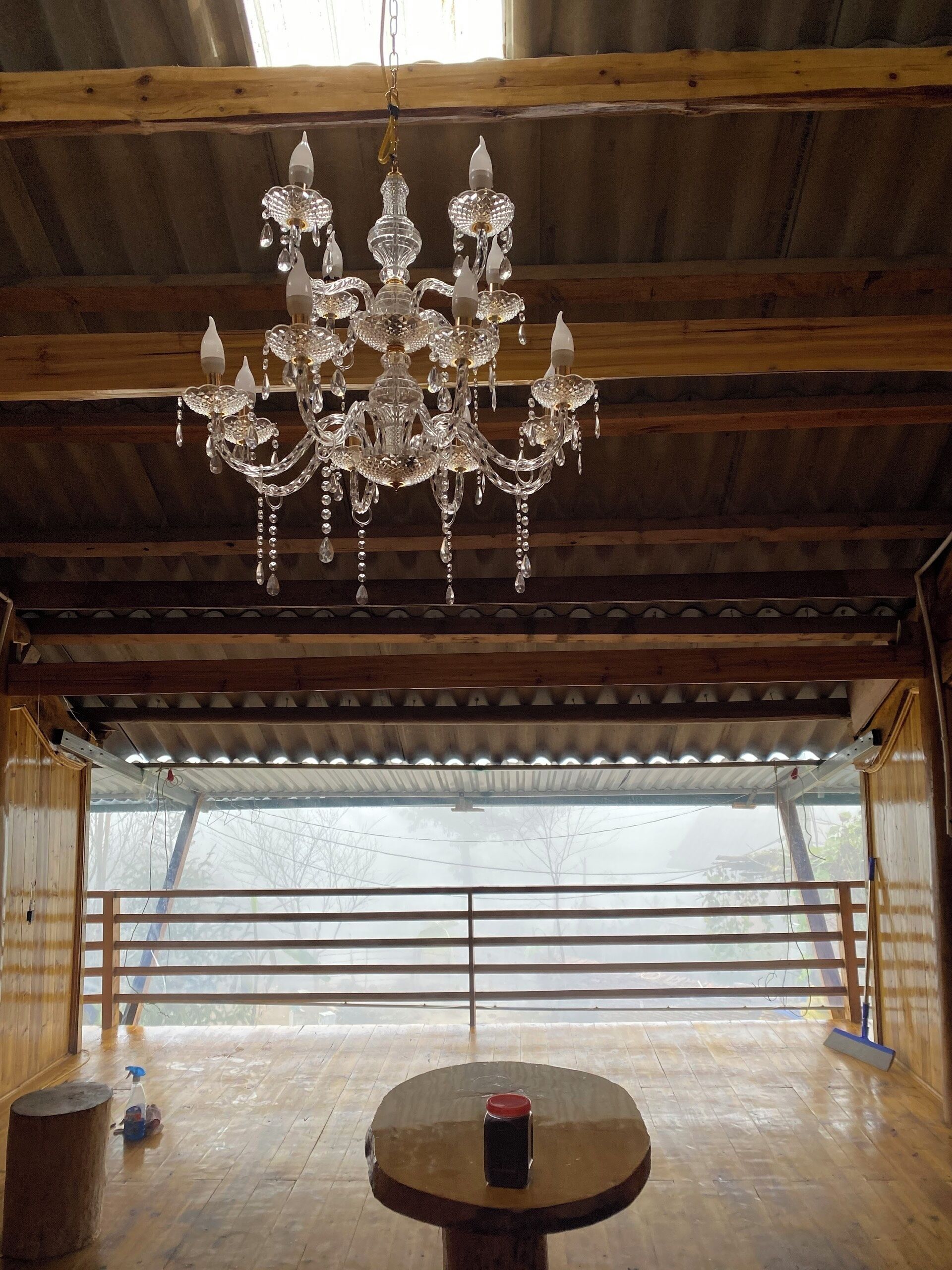 A chandelier hanging from a wooden ceiling in a room with a large window and a round table with a red object on it.