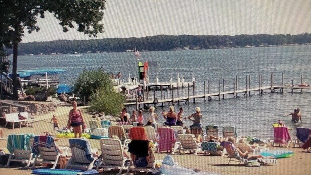 Een strand met ligbedden en parasols, een pier die het water in reikt, en mensen die zwemmen en ontspannen.