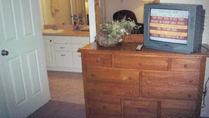 Een houten dressoir met een televisie waarop een nieuwskanaal draait. Een potplant staat op het dressoir, en een spiegel hangt aan de muur.