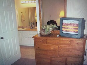 Une commode en bois sur laquelle est posée une télévision diffusant une chaîne d’information. Une plante en pot se trouve sur la commode, et un miroir est accroché au mur.
