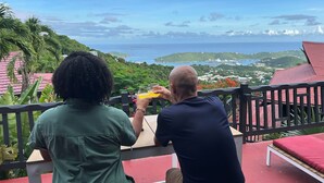 Deux personnes sont assises à une table, devant laquelle s’étend un paysage offrant une vue sur la mer et la verdure.