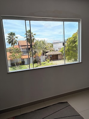 A room with a large window offering a view of a residential area with houses, palm trees, and a clear sky. - Spacious and comfortable apartment in a prime neighborhood in the city of Porto Seguro, Bahia. (Porto Seguro)