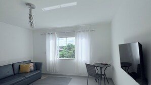 A modern living room with a sofa, a small table, a chair, a flat-screen TV mounted on the wall, and a window with curtains. - Spacious and comfortable apartment in a prime neighborhood in the city of Porto Seguro, Bahia. (Porto Seguro)