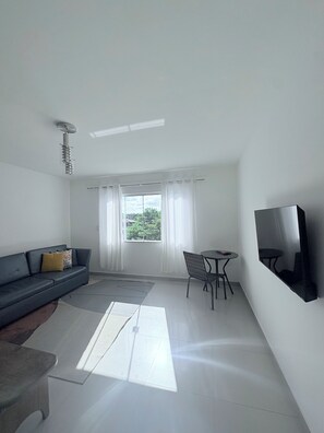 A modern living room with a sofa, a small table, a chair, a flat-screen TV mounted on the wall, and a window with curtains. - Spacious and comfortable apartment in a prime neighborhood in the city of Porto Seguro, Bahia. (Porto Seguro)