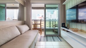 A modern living room with a sofa, a TV, and a balcony with a view of buildings. - Ahead Ponta Negra #602 - Apartment by Carpediem (Natal)