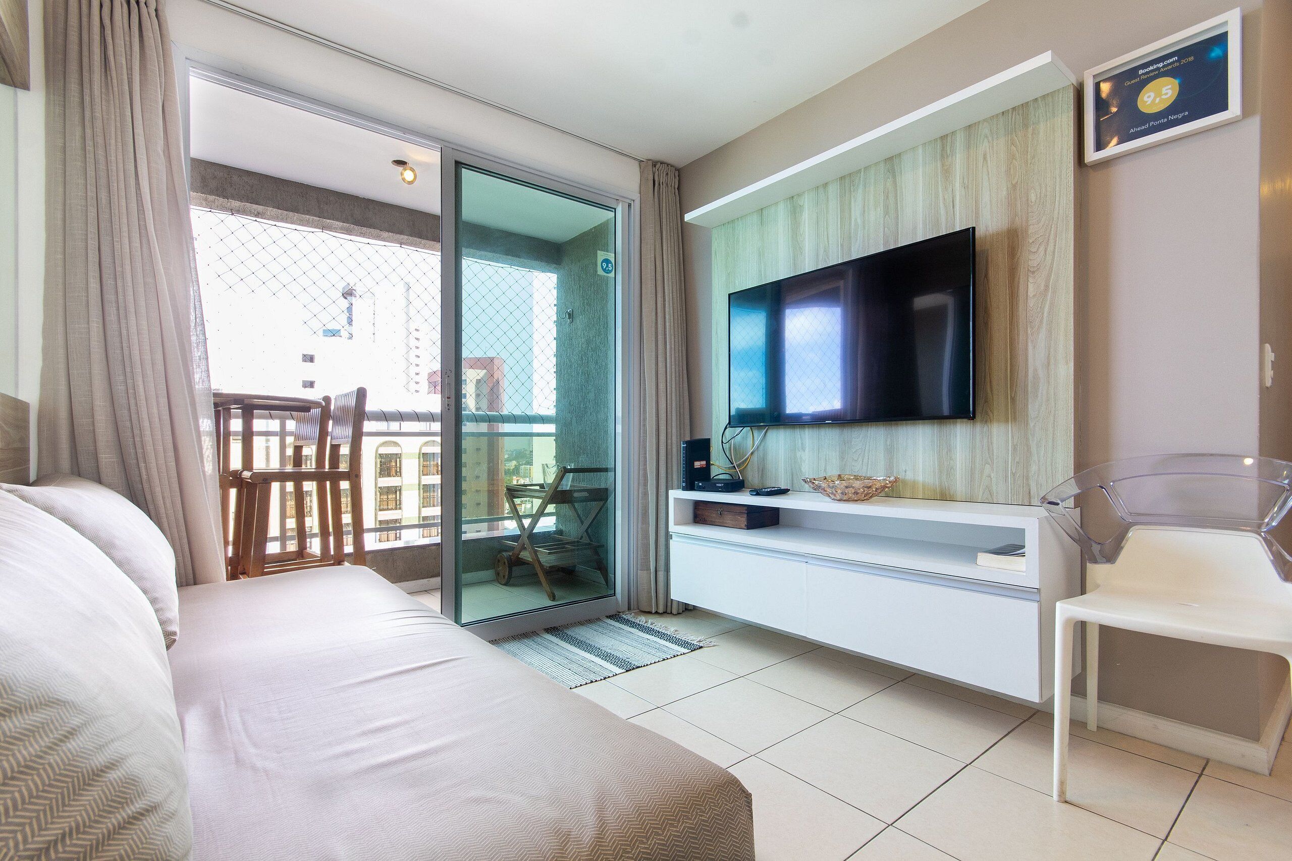 Ein modernes Hotelzimmer mit einem großen Bett, einem an der Wand montierten Fernseher, einer Glastür zu einem Balkon und einem weißen Schminktisch.