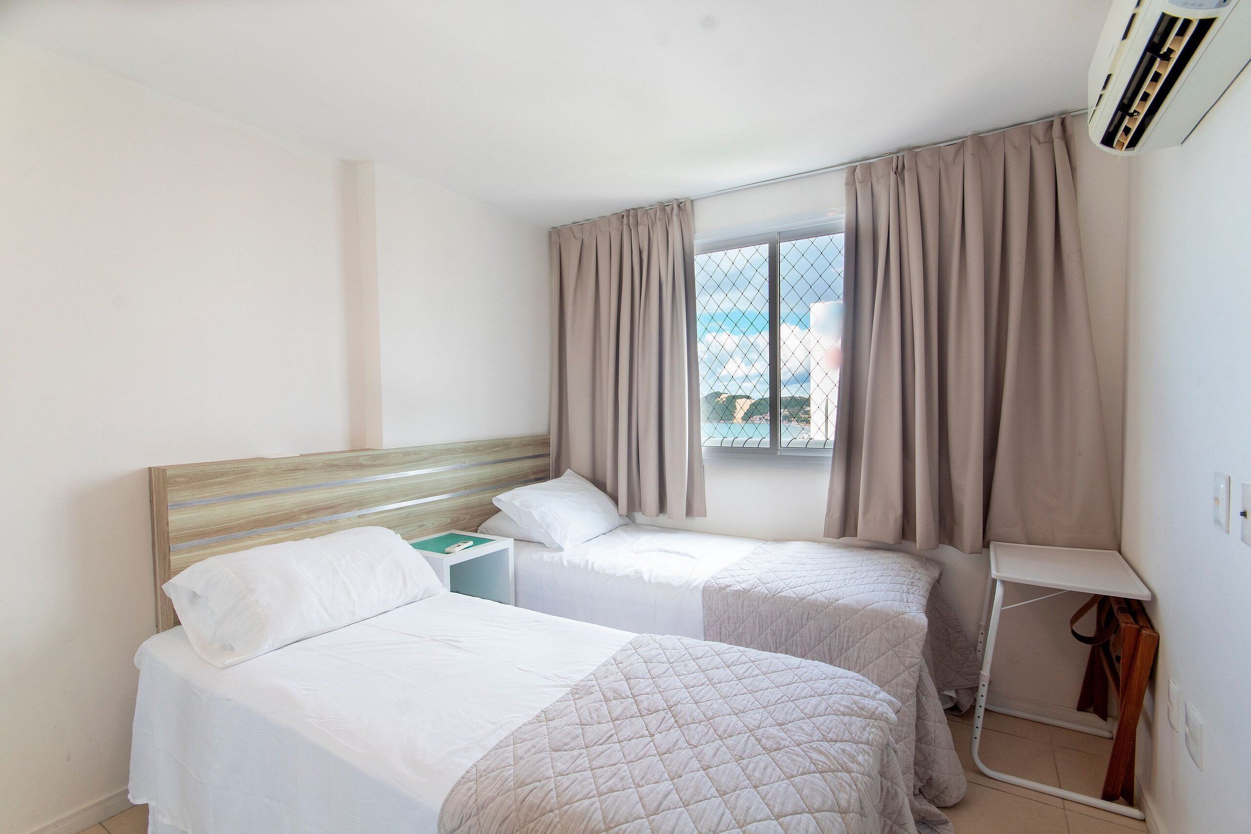 A hotel room with two single beds, a small table, a window with curtains, and an air conditioning unit.