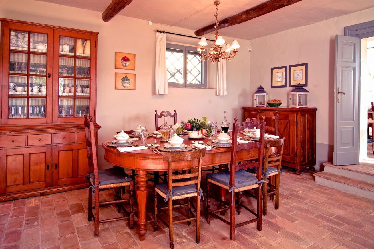 A dining room with a wooden table set for four, surrounded by chairs. A wooden cabinet with glass doors is to the left, and a chandelier hangs from the ceiling.