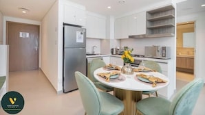 A modern kitchen with white cabinets, a stainless steel refrigerator, and a dining area with a round table and green chairs. - Vogue | Downtown with Direct Access to Dubai Mall (Dubai)