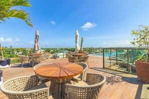 A rooftop terrace with wicker furniture, a pool, and a glass railing overlooking a cityscape. - Rooftop Orla Tamandare #205 - Apto by Carpediem (Tamandaré)