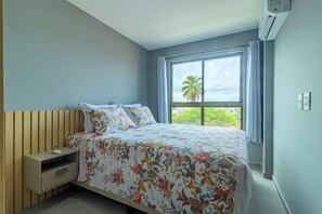 A bedroom with a bed, a nightstand, a window with curtains, and an air conditioning unit. - Rooftop Orla Tamandare #205 - Apto by Carpediem (Tamandaré)